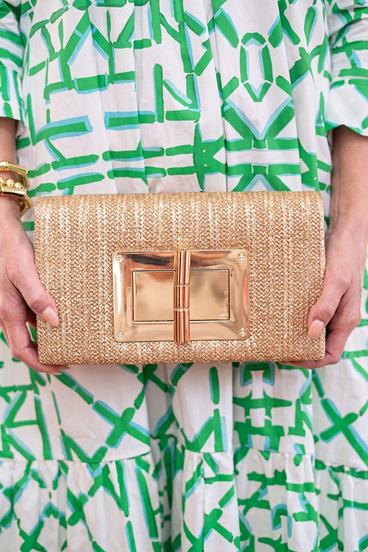 The Gold Lillian Straw Clutch held in both hands against a green and white patterned dress.