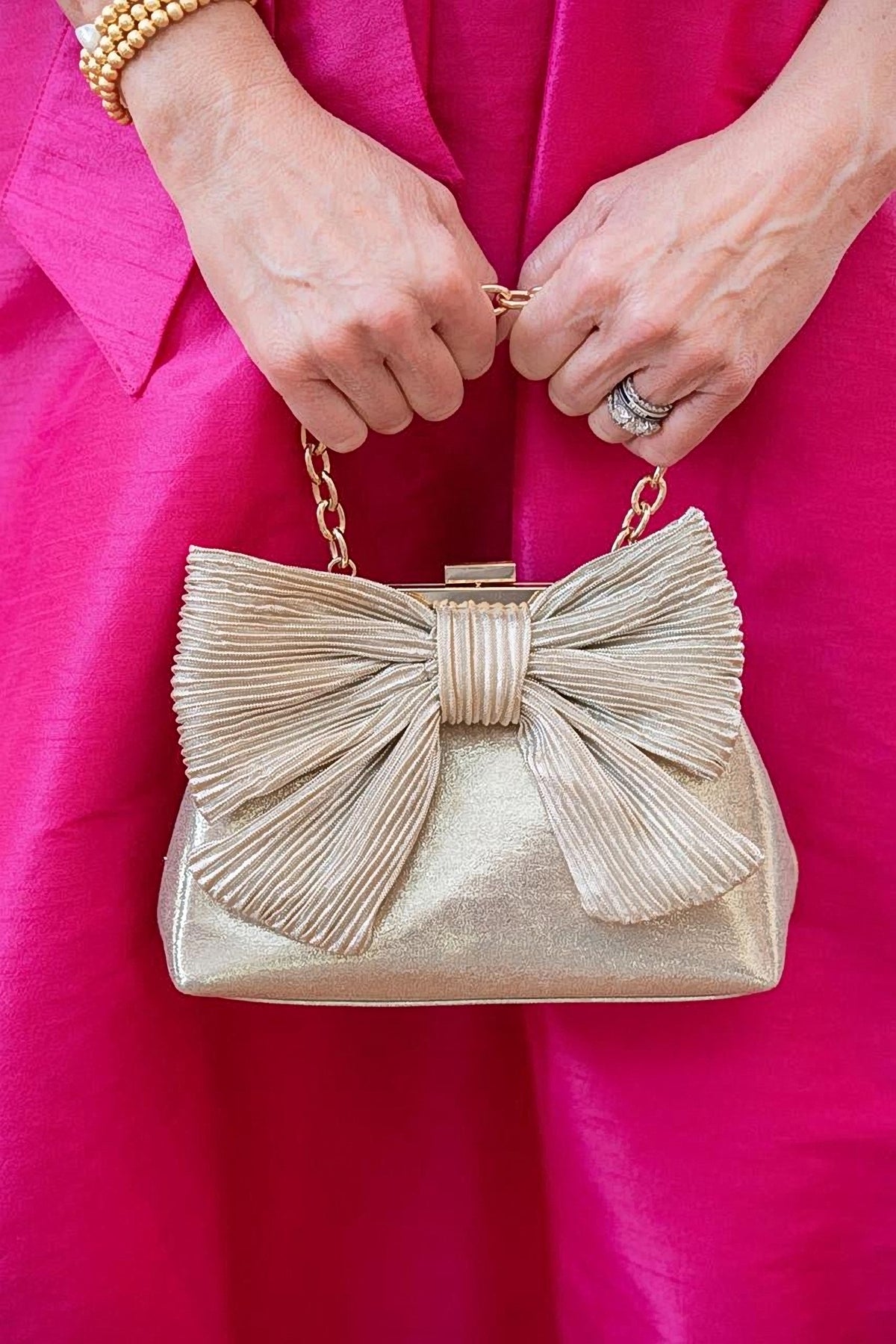 The gold Samantha Bag held by its short chain in front of a hot pink dress.