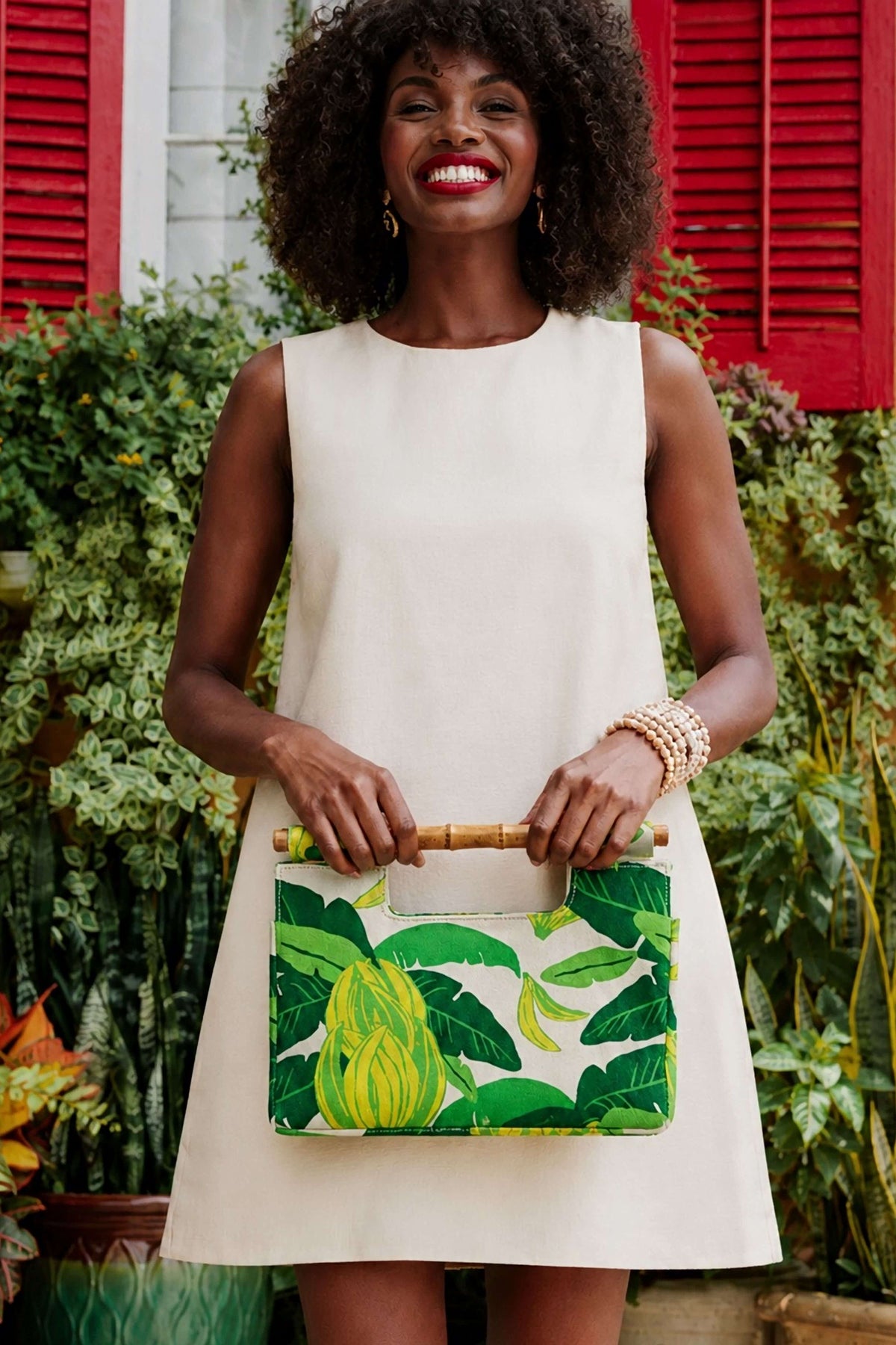 The Banana Bamboo Clutch held by its bamboo handles with a cream shift dress in front of a red shuttered house.