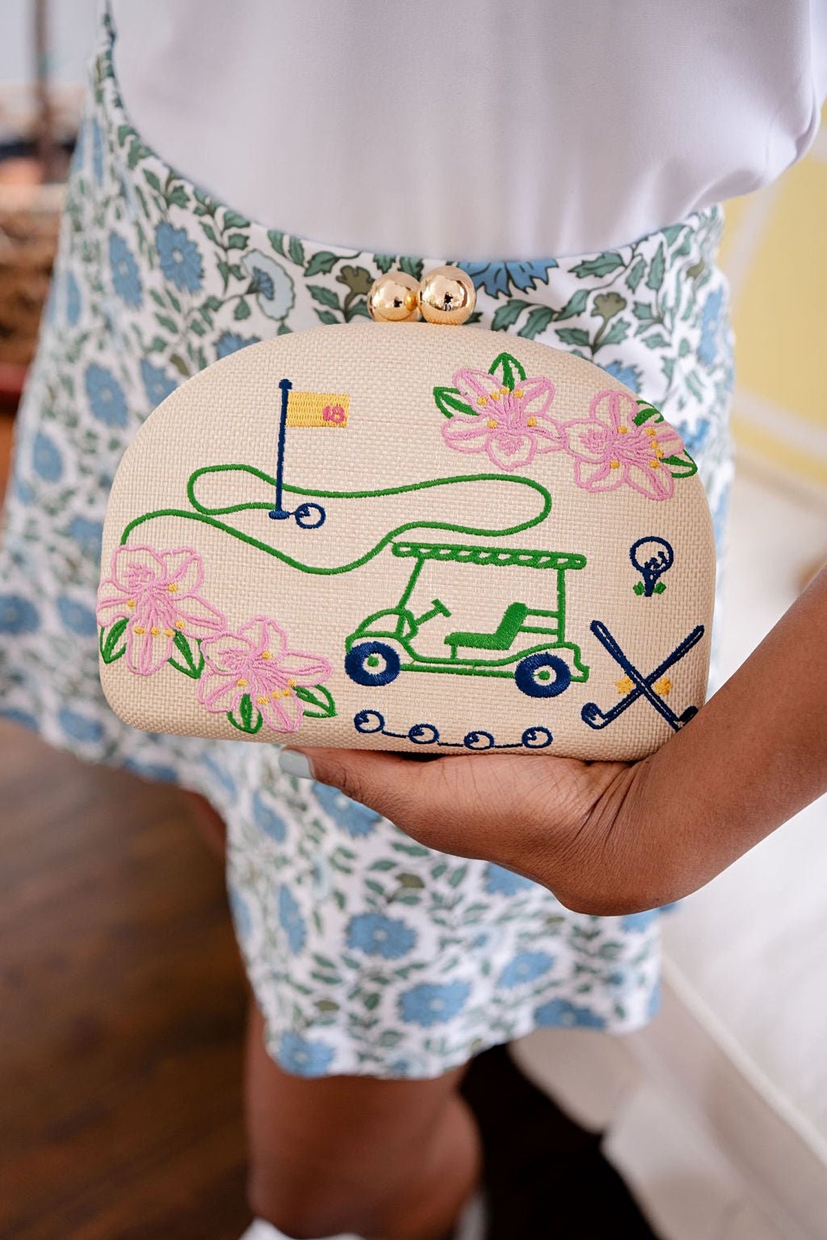 The Golf Embroidered Clutch held under one hand against a blue floral skirt and white top.