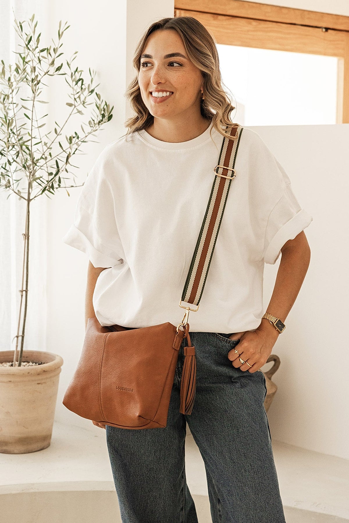 The Daisy Stripe Tan Crossbody Bag worn at the hip on its striped webbing strap with a white tee and jeans.