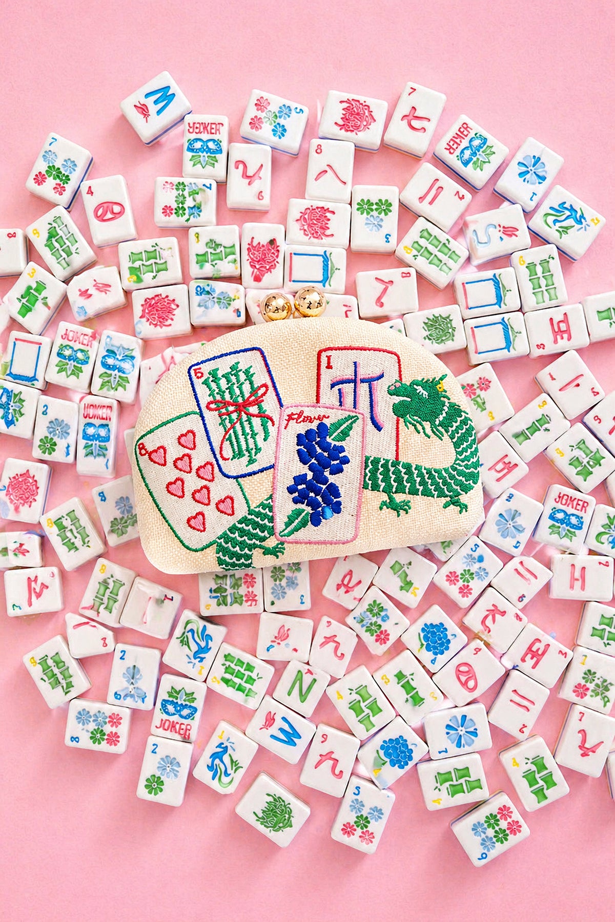 The Mahjong Embroidered Clutch laid on a pink tabletop scattered with mahjong tiles.