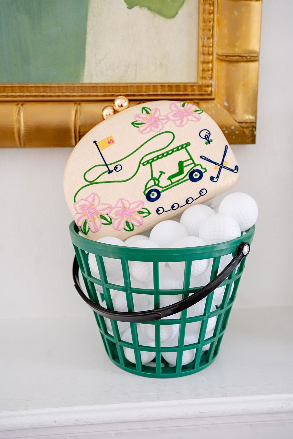 The Golf Embroidered Clutch propped on a green basket of white golf balls in front of a gold framed painting.