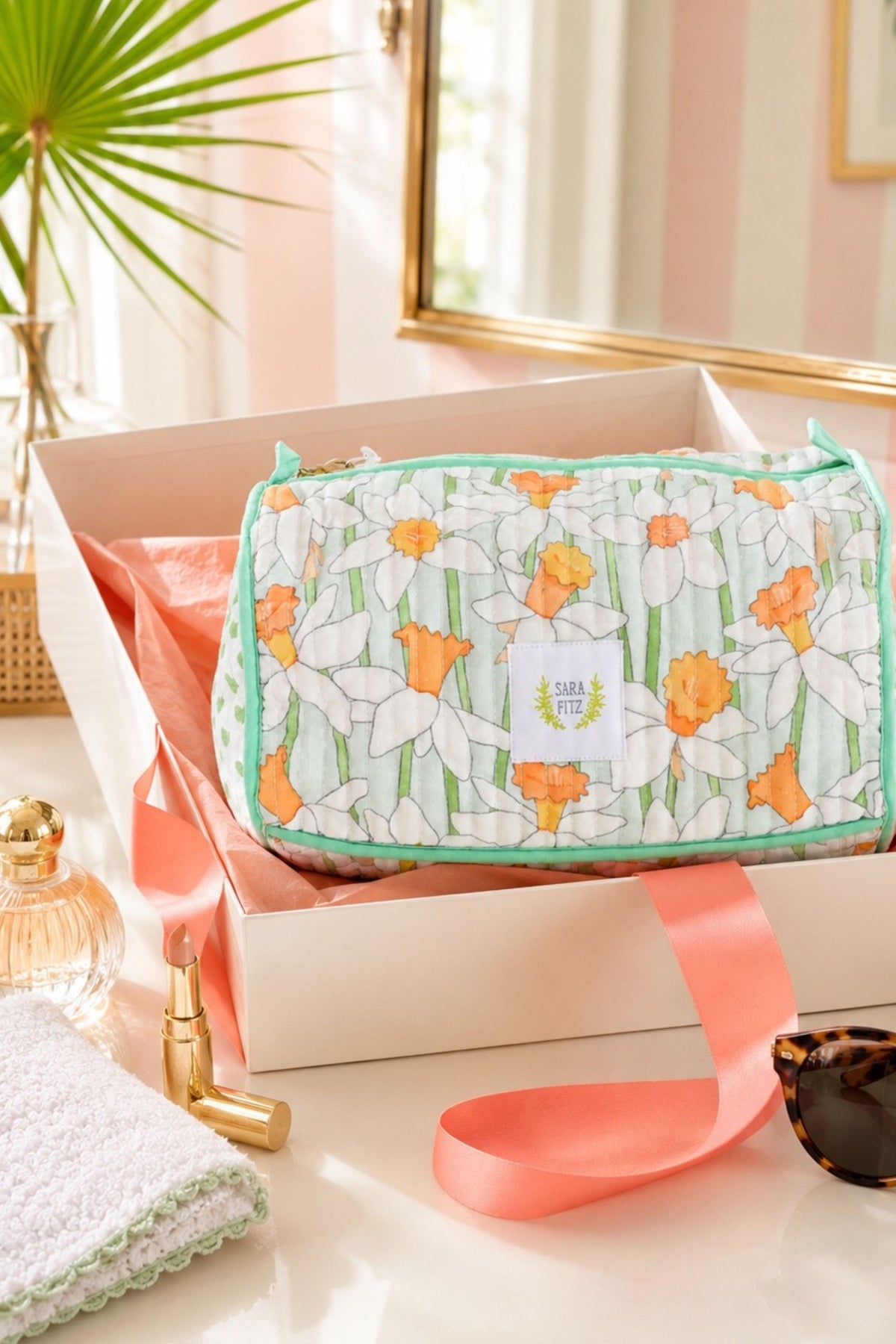 A cosmetic pouch with a daffodil floral print in white, green, and orange, presented inside a white gift box with pink ribbon. The pouch features a white label with the brand name 'Sunny Day'.