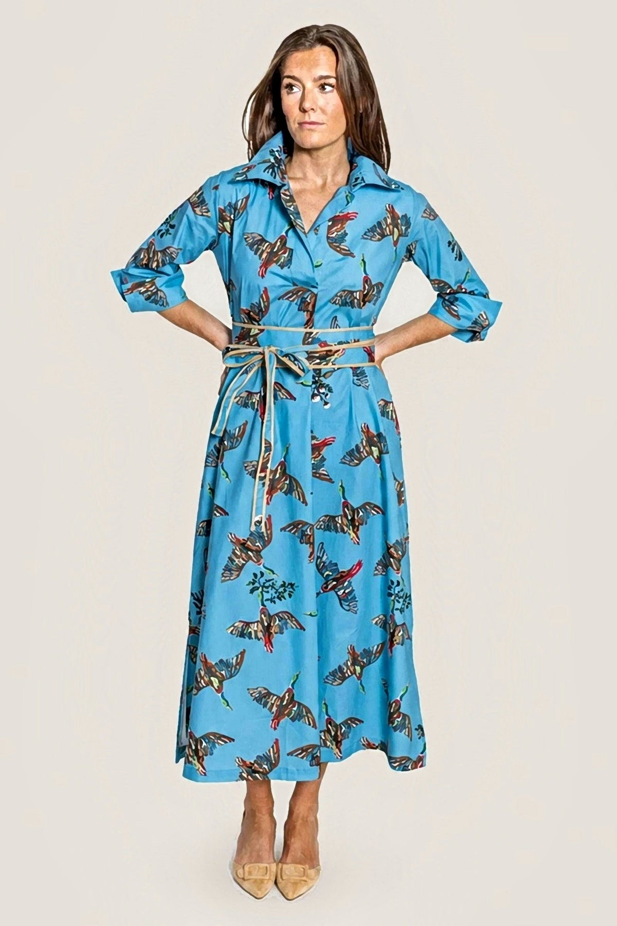 A woman modeling a blue dress with pheasant birds and a tie belt wrapped around her waist high-heeled sandals against a white studio backdrop.
