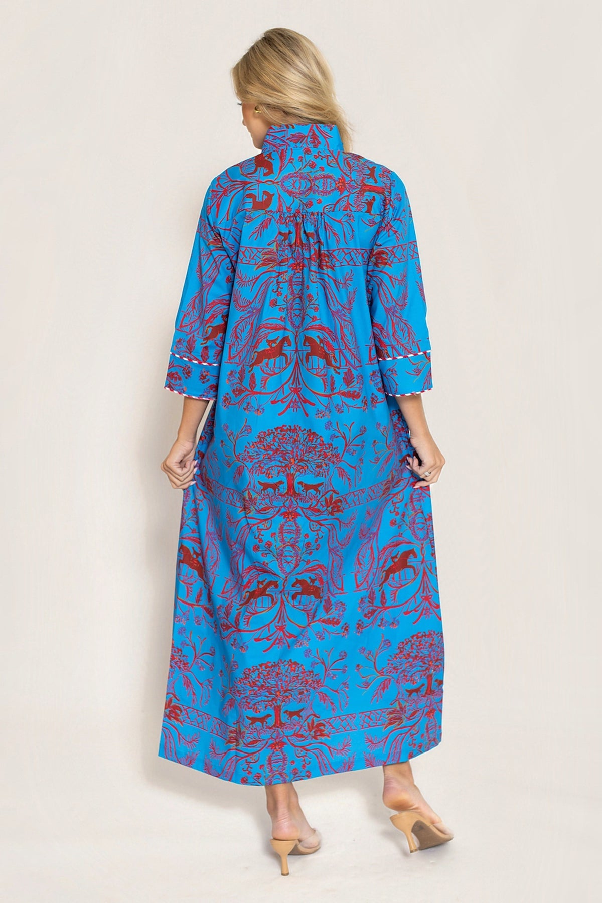 A woman viewed from the back wearing a long blue dress with a red and purple botanical pattern, standing against a plain off-white studio background.