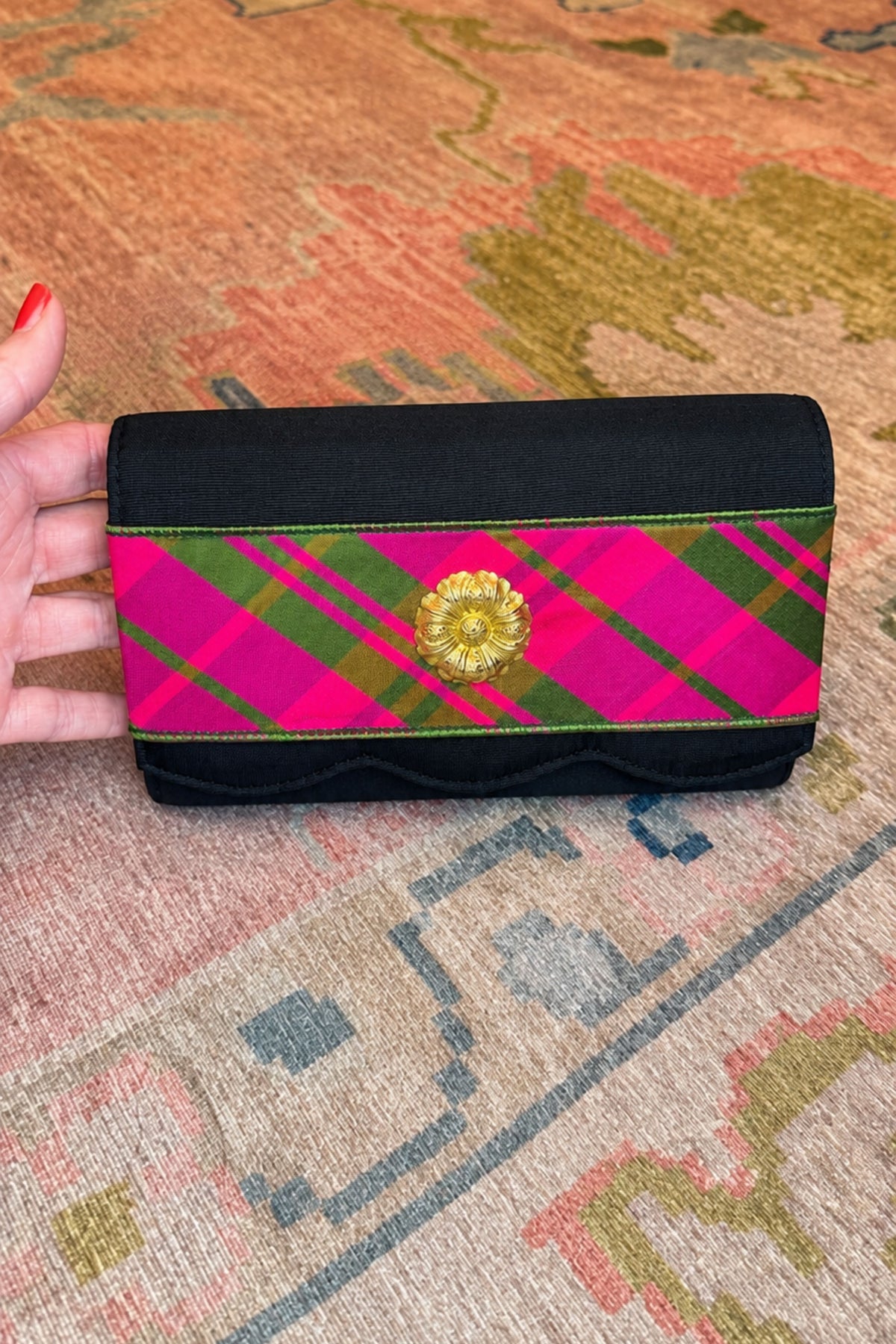 A hand holds a black fabric clutch wallet featuring a vibrant pink and green tartan plaid band across the front with a gold-tone floral embellishment in the center, set against a patterned rug.