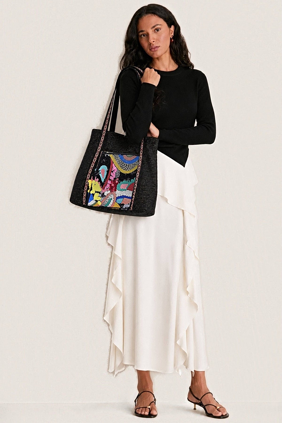 A woman in a black sweater and white ruffled skirt carrying the Black Triple Entry Embellished Shoulder Bag on her shoulder, its beaded front panel facing out.