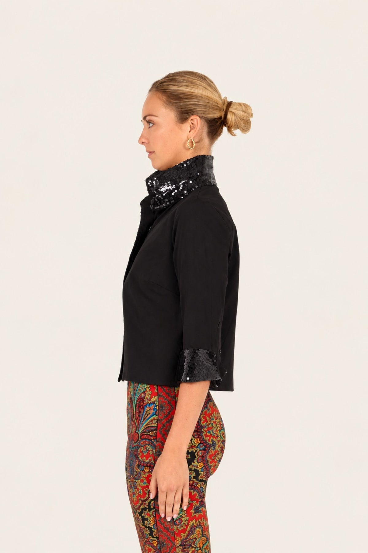 A woman shown in side profile wearing a black cropped jacket with a high black sequined collar and matching black sequined cuffs, paired with red and gold paisley pants, against a plain light background.
