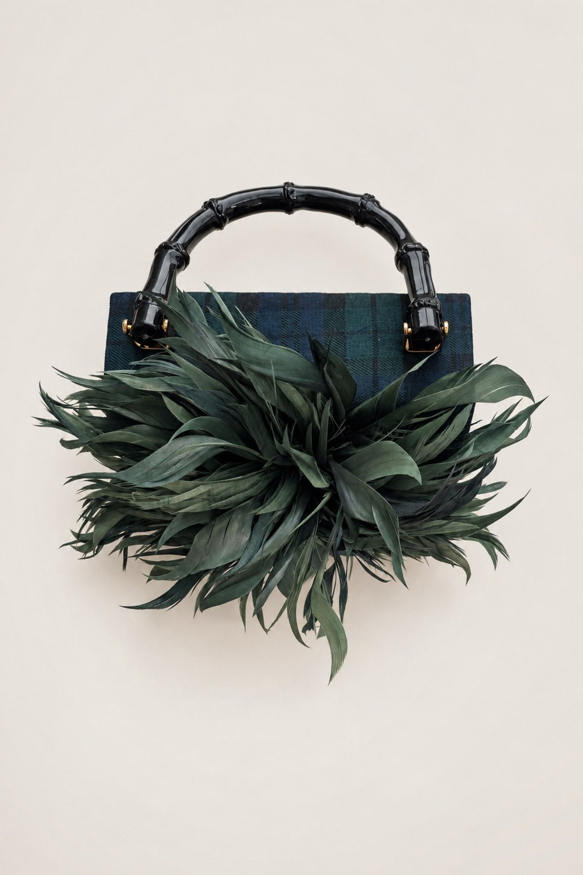 A structured handbag with a dark green plaid body, a glossy black bamboo-style top handle, and a large, voluminous dark green feather or leafy embellishment in front, photographed against a plain warm beige background.