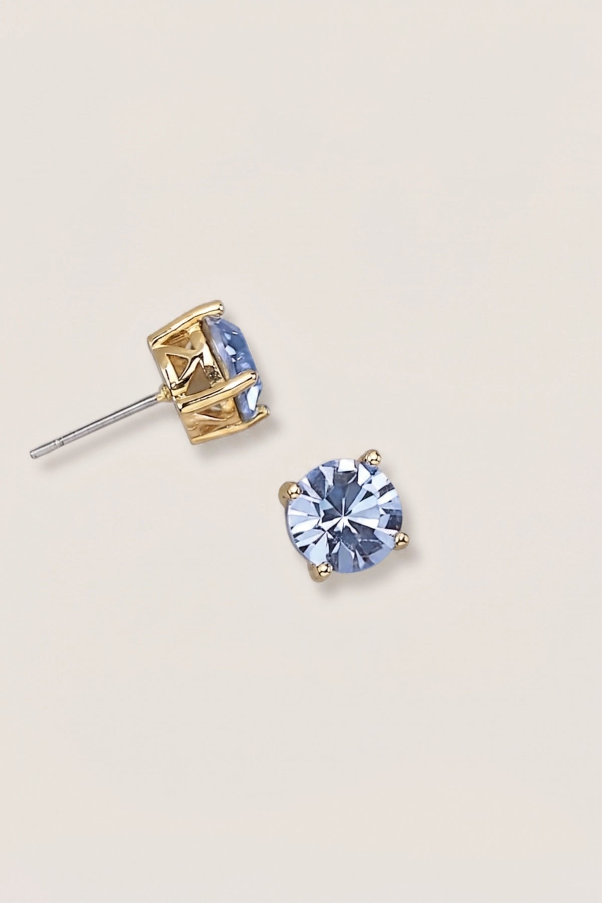 A pair of gold stud earrings with oval light blue gemstones, one shown from the front and the other from the side/back, on a light cream background.