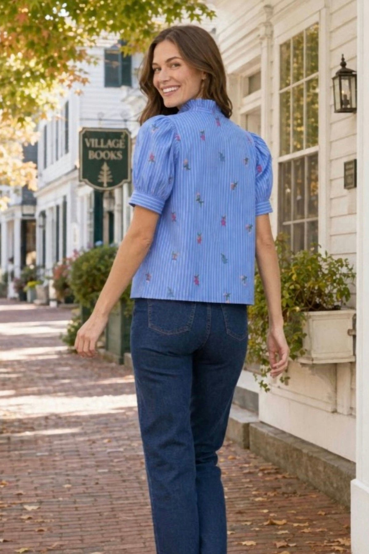 A woman wearing a light blue short-sleeve button-up blouse with vertical stripes and small colorful embroidered motifs, paired with dark blue jeans, walking on a brick street in front of a white building.
