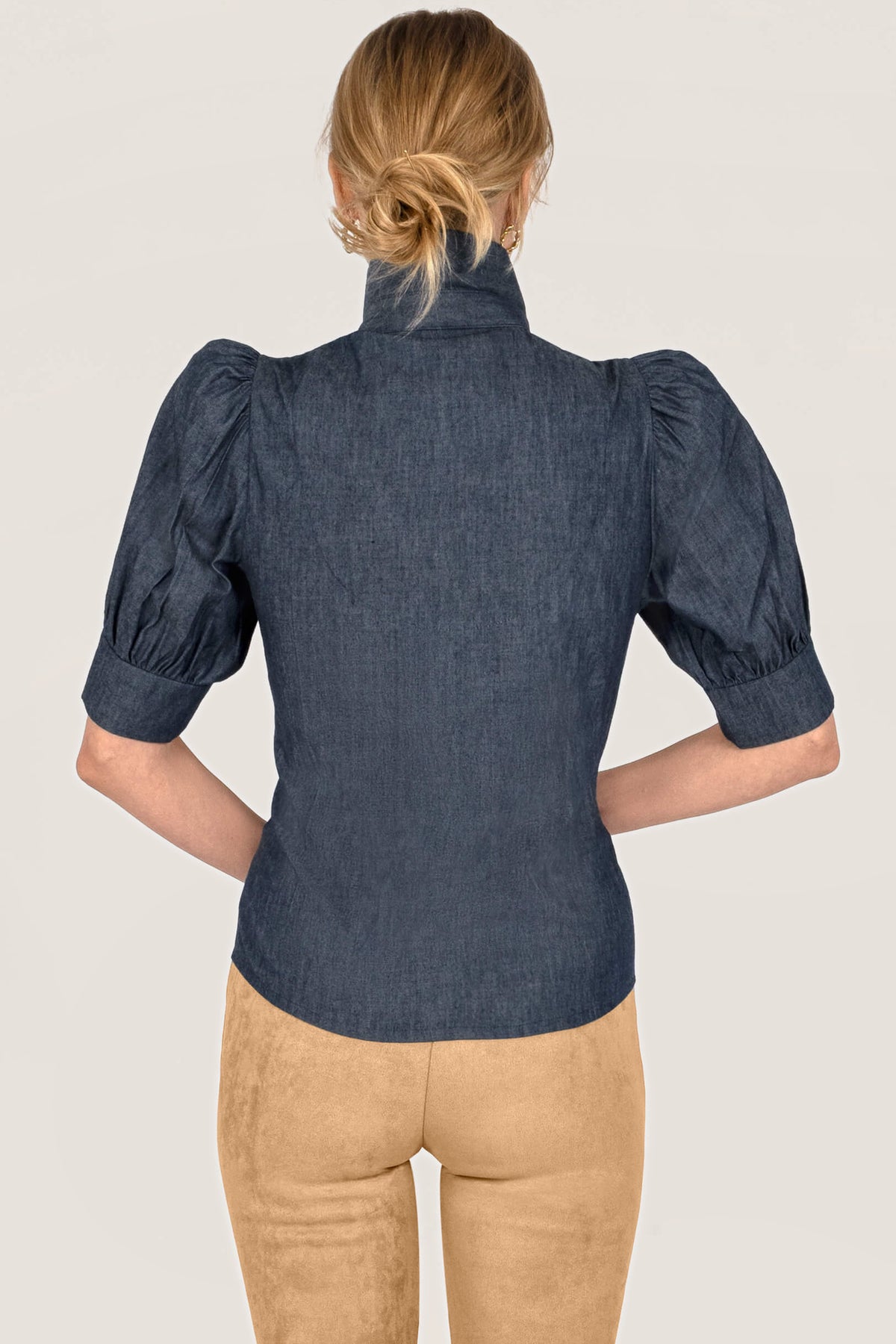 A woman shown from the back wearing a dark blue chambray puff-sleeve blouse and tan pants against a plain off-white background.