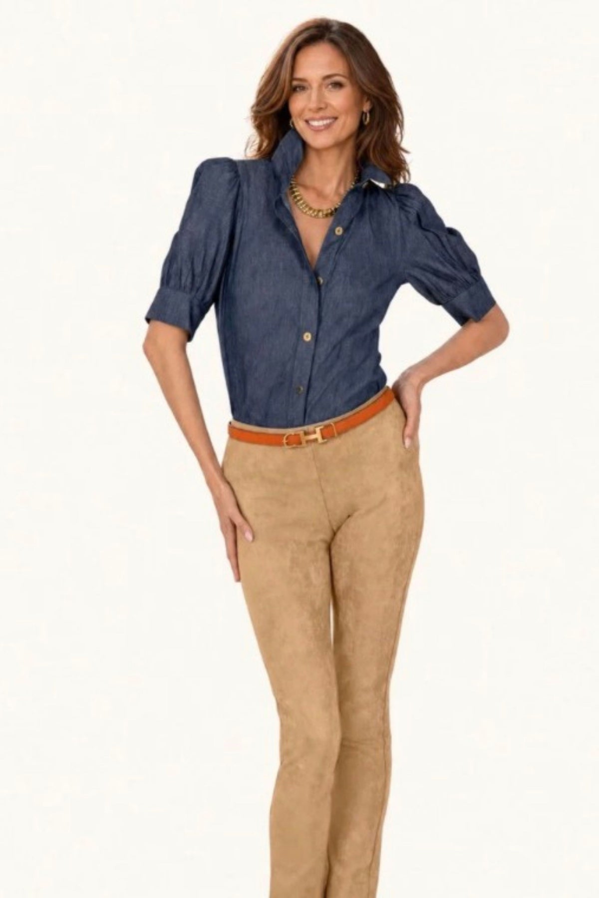 A woman modeling a dark blue chambray button-down shirt with puff sleeves and tan suede pants, styled with a brown belt, against a plain white background.
