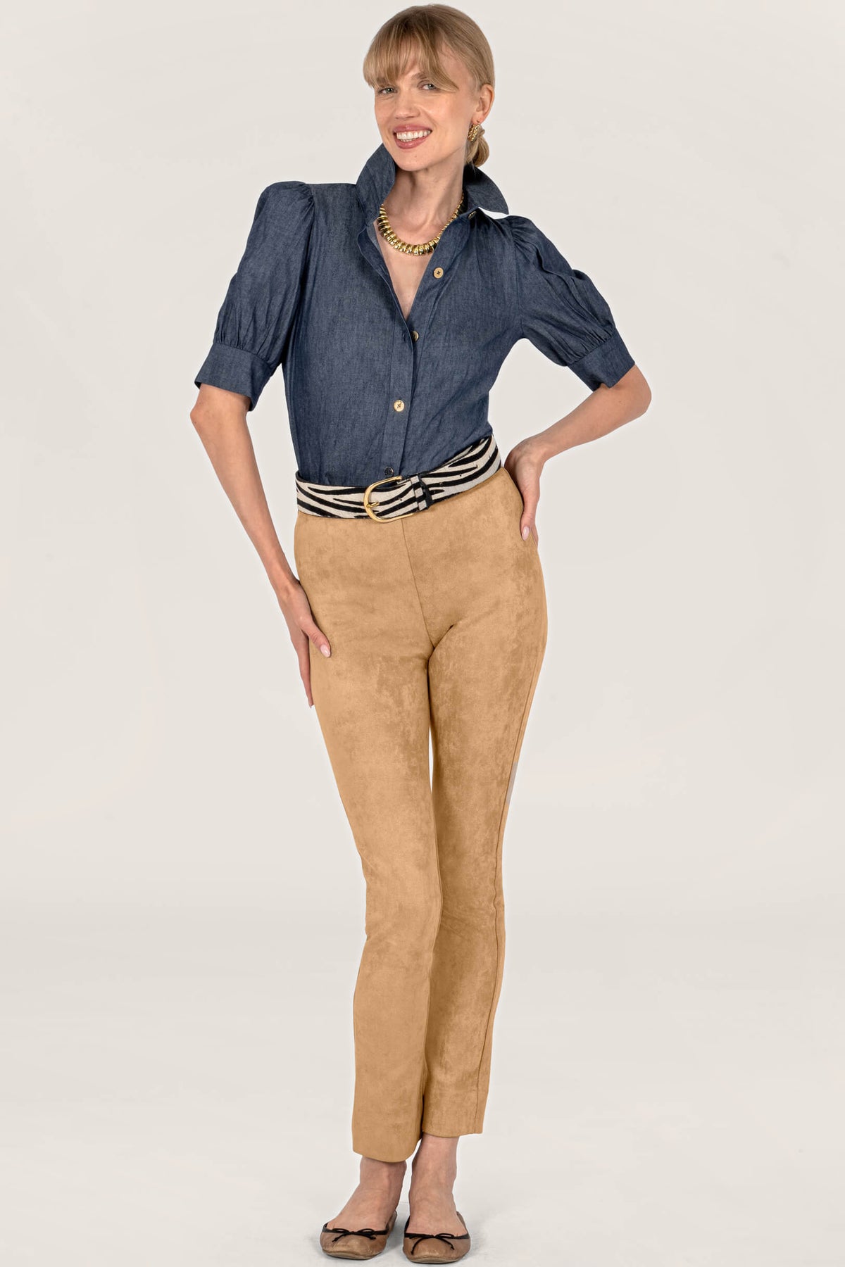 A woman modeling a dark blue chambray button-down shirt with puff sleeves and tan suede pants, styled with a brown belt, against a plain white background.
