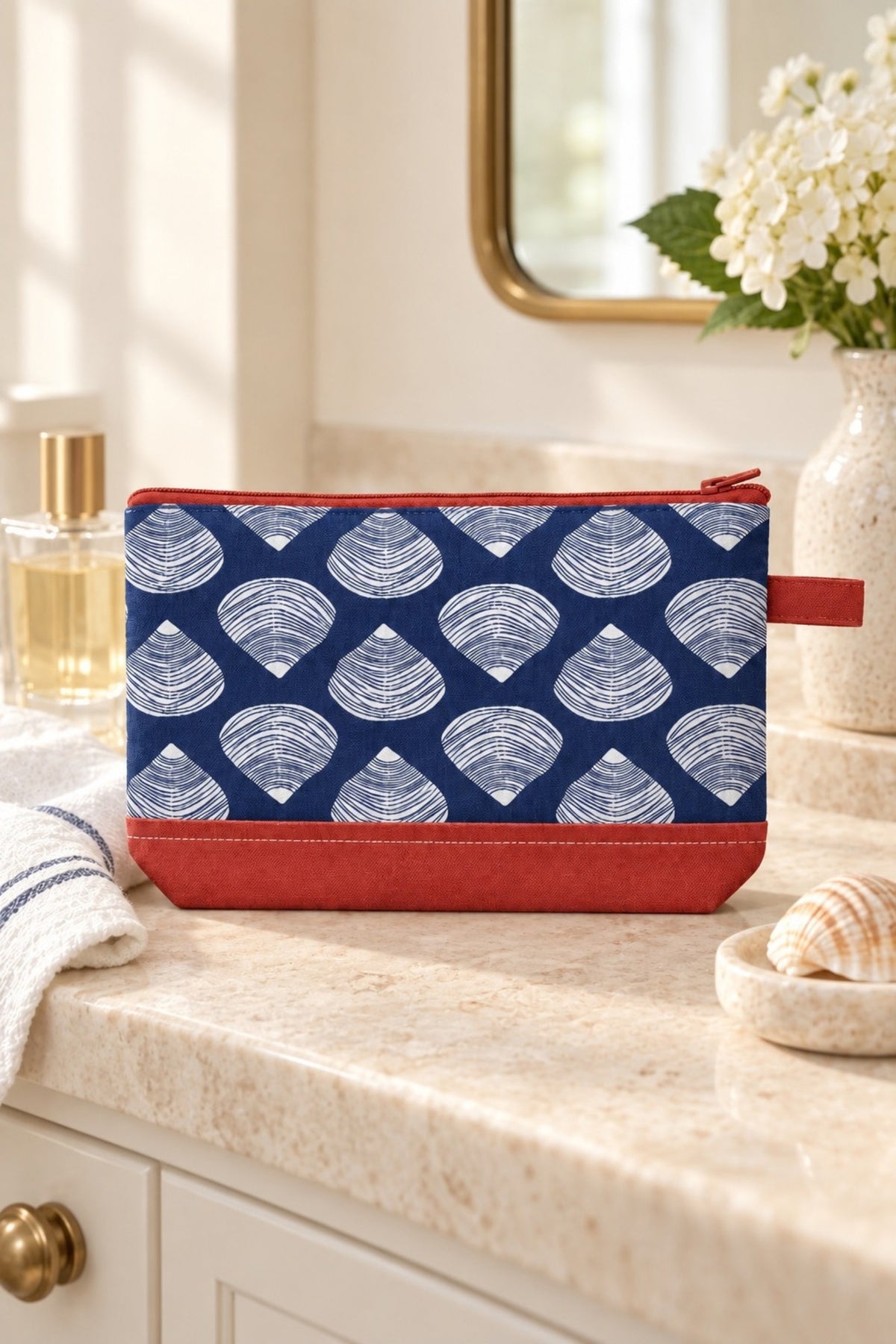 The Clamshell cosmetic bag on a bathroom vanity beside a mirror and white flowers.
