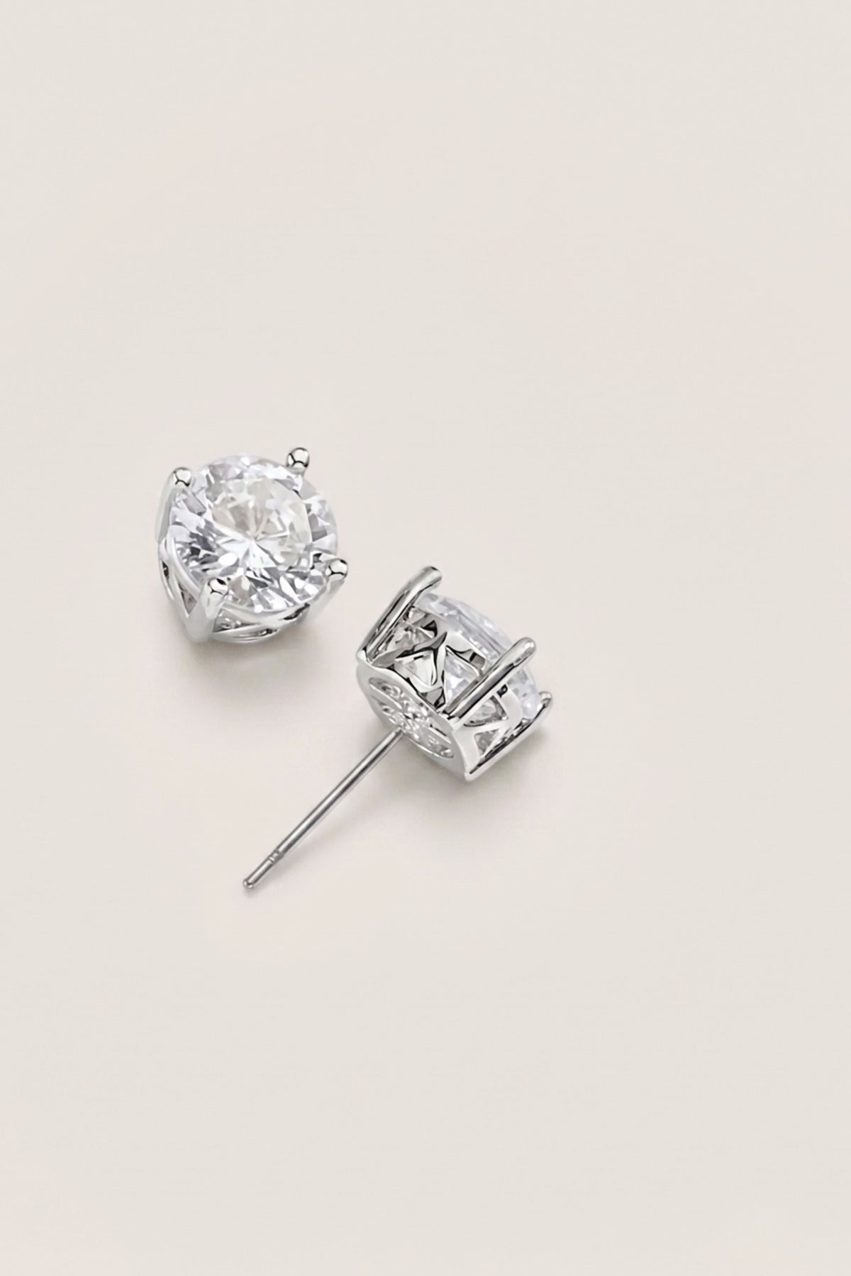 A pair of silver stud earrings with clear gemstones, one shown from the front and the other from the side, on a light beige background.