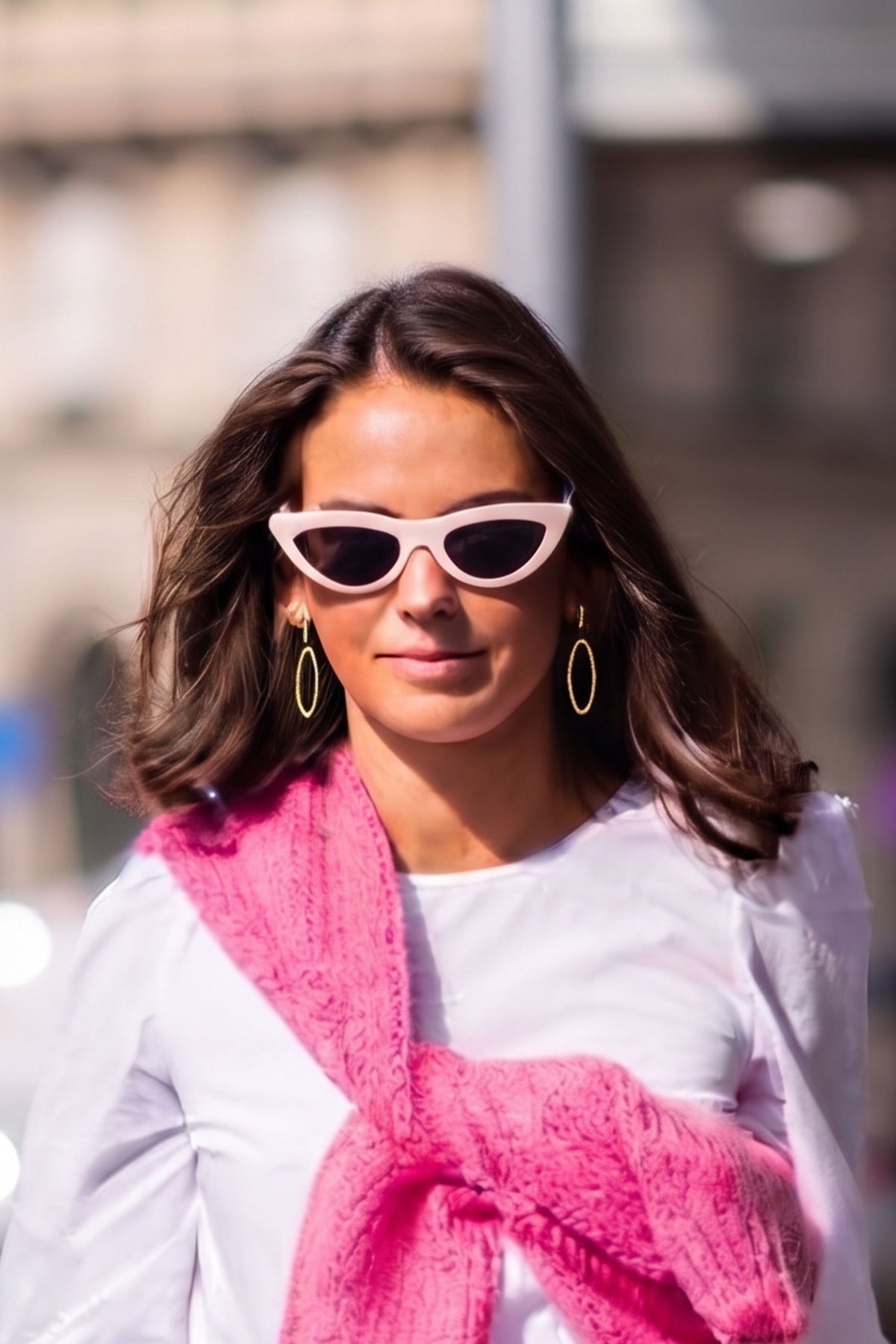 A woman wearing white cat-eye sunglasses with dark lenses, a white top, and a bright pink knitted scarf, photographed outdoors with a blurred city background.