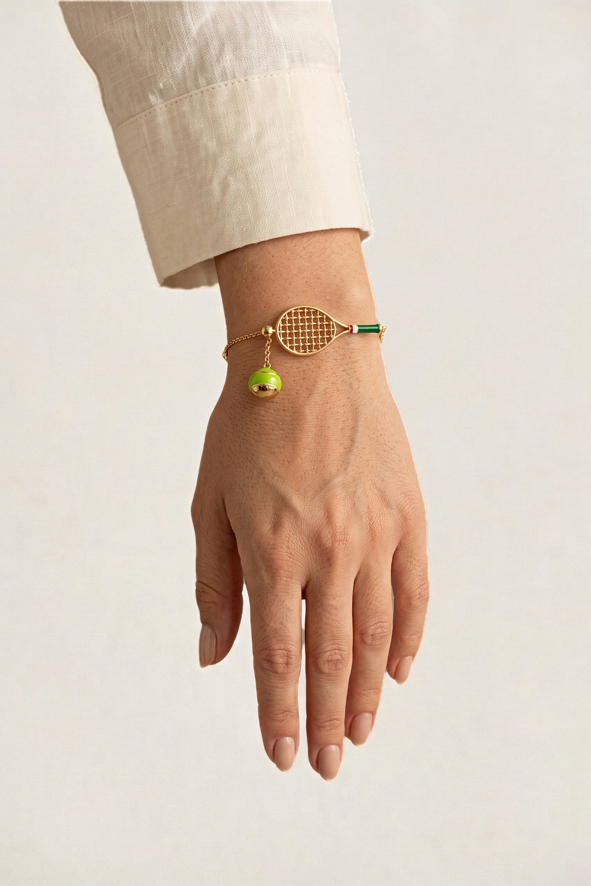 Country Club Tennis Bracelet worn on the wrist with a cream linen sleeve, showing the gold racket charm and the green and gold tennis ball charm.