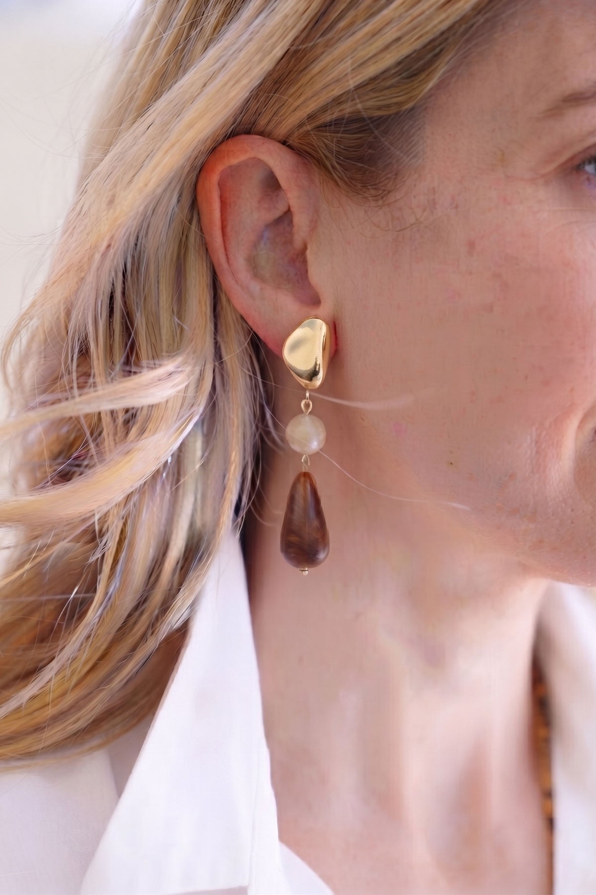 A close-up of a woman's ear wearing a dangling earring with a gold top, a small white bead, and a dark brown teardrop stone.