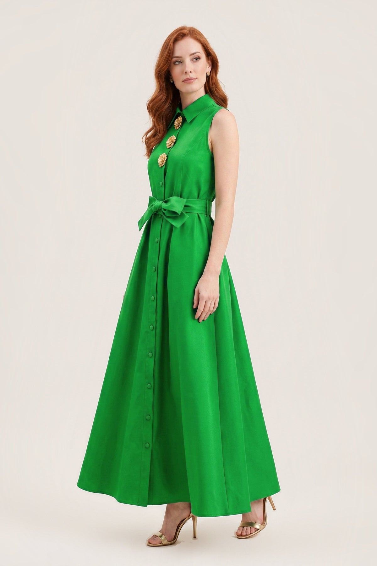 A woman modeling a sleeveless emerald green maxi dress with a high collar, gold decorative buttons on the chest, a matching green waist tie, and front button detail, photographed against a plain light beige studio background.