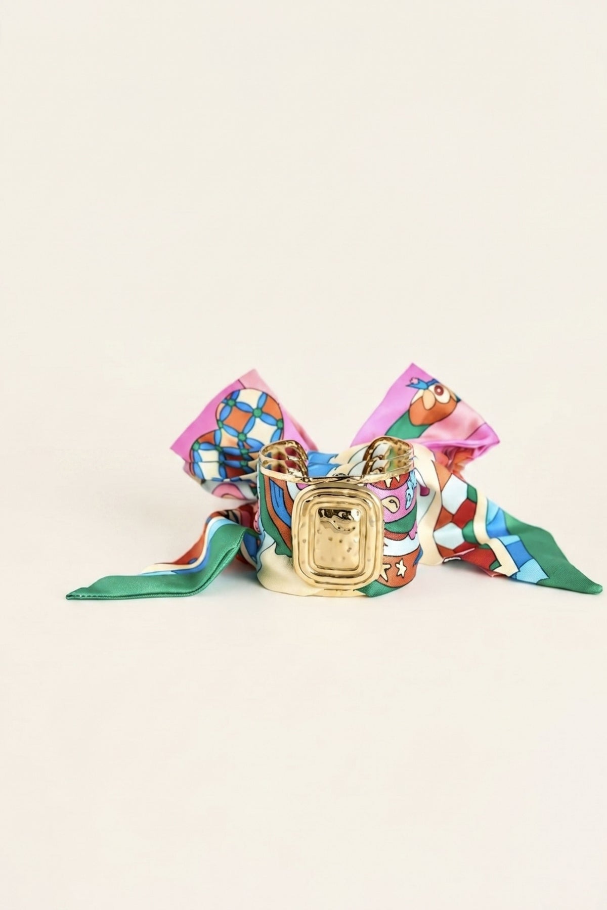 A colorful patterned scarf styled as a bow with a small gold-toned jewelry box placed in the center, photographed on a plain light beige background.