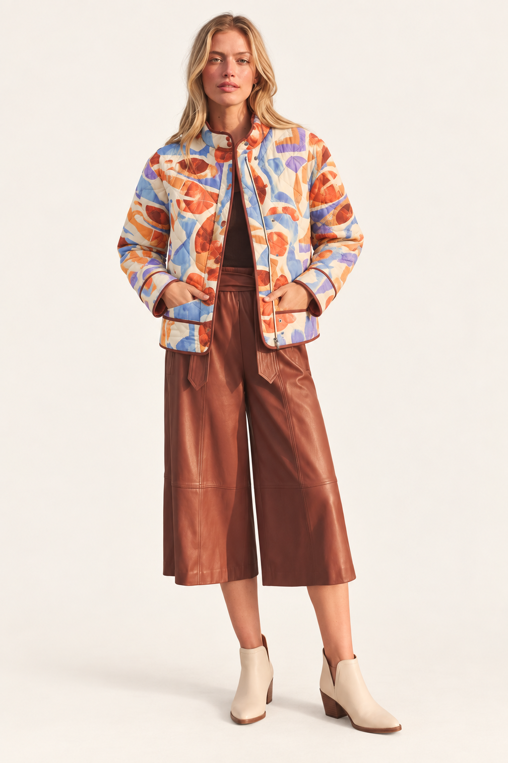 A woman modeling a colorful abstract print bomber jacket, paired with brown wide-leg faux-leather cropped pants and cream ankle boots, against a plain off-white studio background.