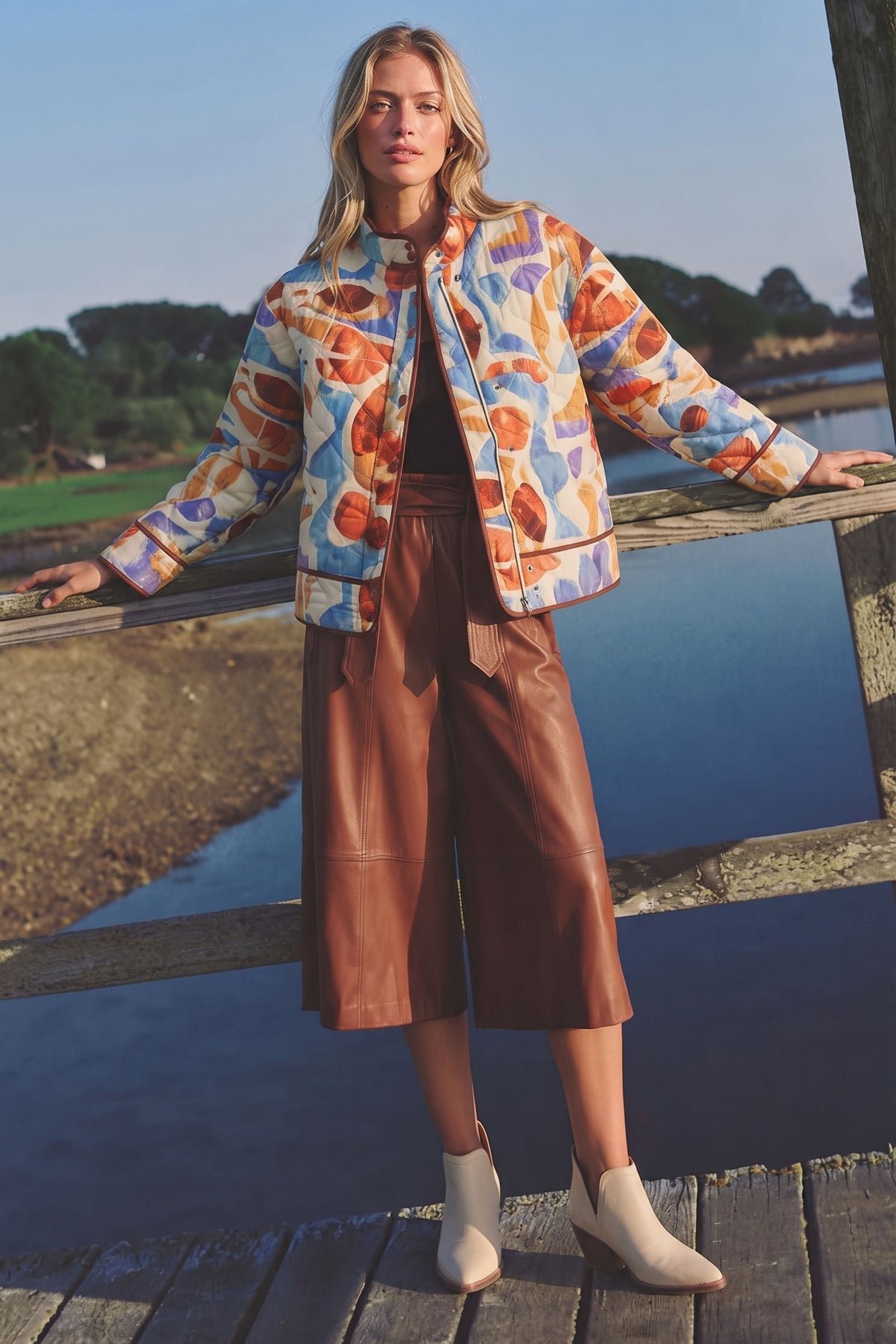 A woman stands on a wooden bridge outdoors wearing a colorful abstract print bomber jacket, brown cropped leather pants, and white ankle boots, with water and trees in the background.