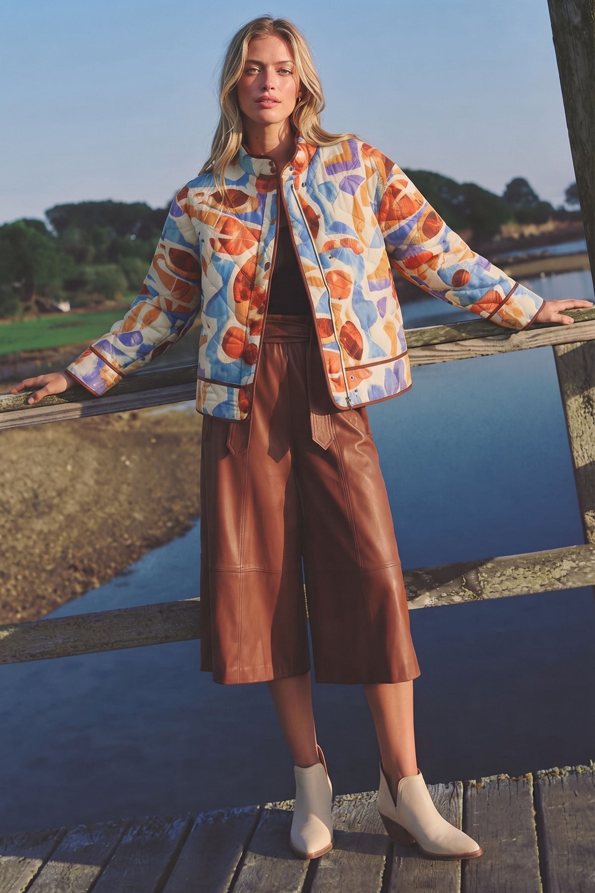 A woman stands on a wooden bridge outdoors wearing a colorful abstract print bomber jacket, brown cropped leather pants, and white ankle boots, with water and trees in the background.