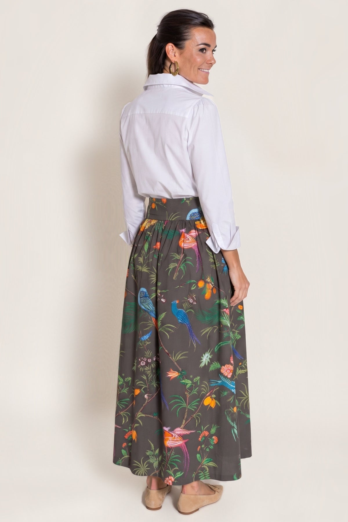 A woman shown from the back wearing a white button-up shirt and a dark chocolate gray maxi skirt with a colorful tropical bird and floral print, standing against a plain beige studio background.