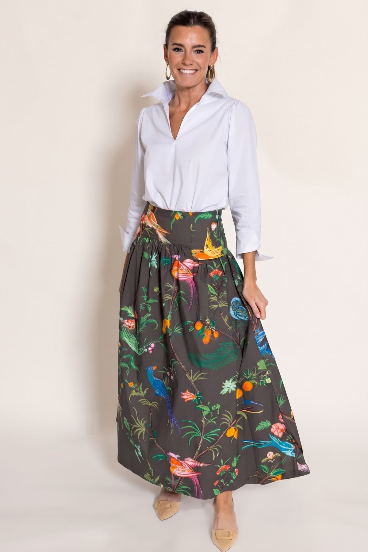 A woman modeling a dark chocolate maxi skirt with a colorful tropical bird and floral print, paired with a white button-down shirt, against a plain beige studio background.