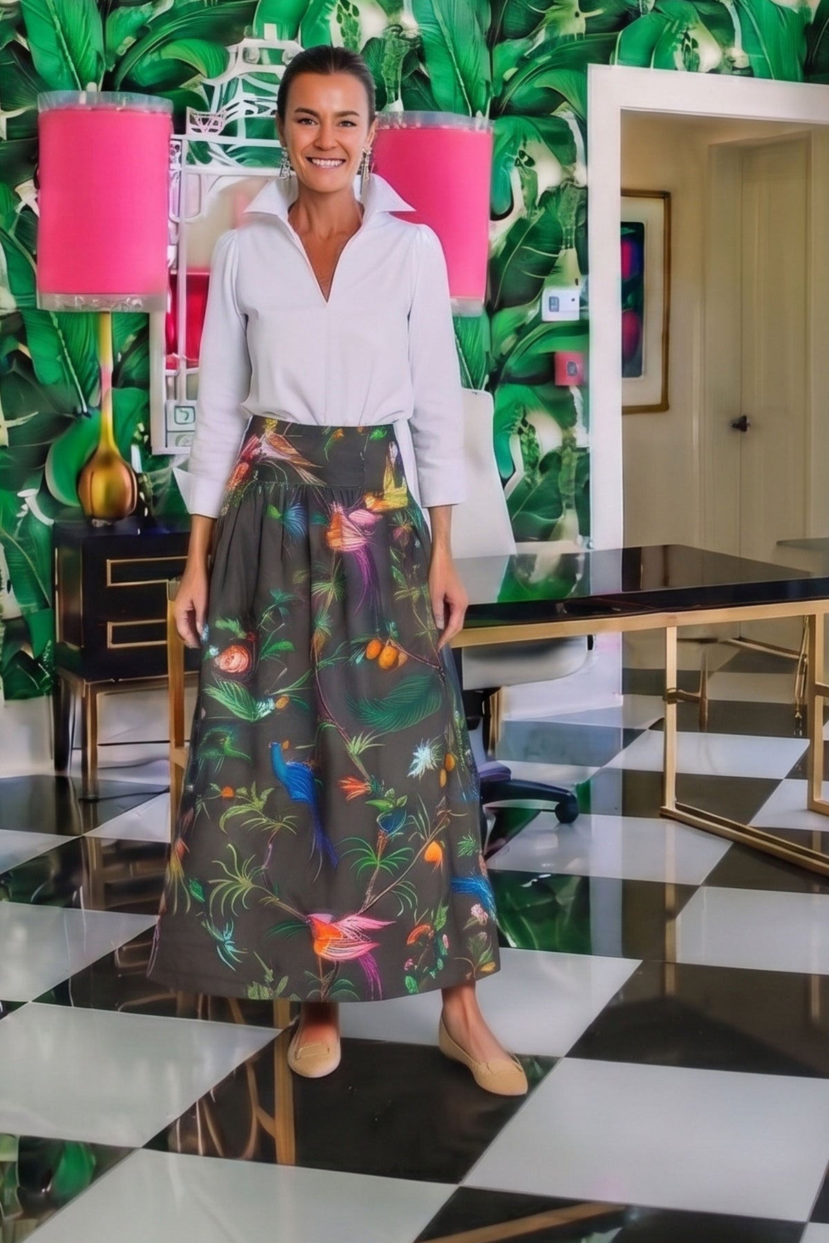 A woman stands in a stylish room wearing a white collared blouse tucked into a high-waisted, full-length skirt with a colorful tropical bird and floral print on a dark gray background. The room features bold green tropical wallpaper, pink lamps, a black-and-white checkered floor, and a desk.