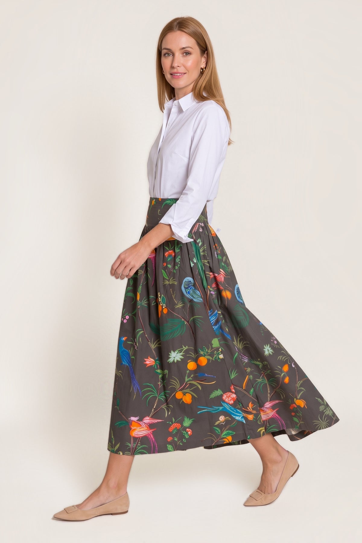A woman walking in the Feather Frolic Sadie drop-waist maxi skirt, a charcoal skirt printed with colourful birds, oranges and botanicals, worn with a white button-down shirt, on a warm neutral studio background.