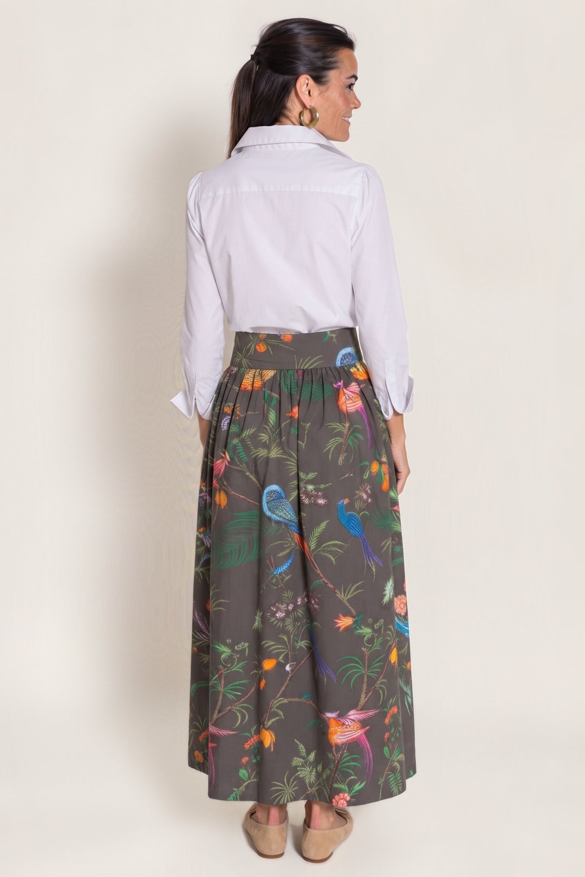 A woman shown from the back wearing a white long-sleeve button-up shirt and a high-waisted, wide-leg pleated skirt with a colorful tropical bird and floral print on a dark chocolate gray background. She is wearing beige flat shoes against a plain beige studio background.