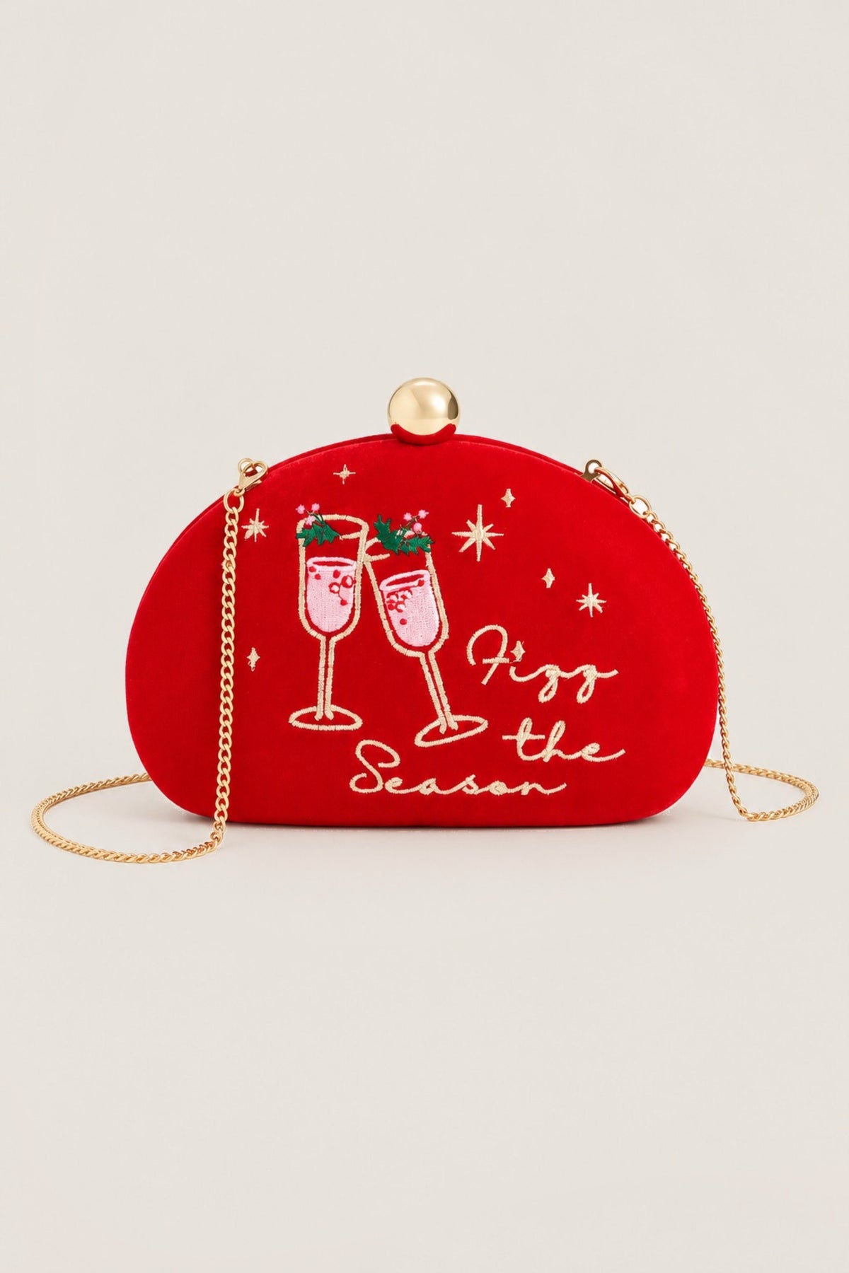 A red velvet crescent-shaped clutch bag with a gold-tone chain strap and top clasp. The front features white embroidered champagne glasses, festive starbursts, and the text 'Fizz the Season'.