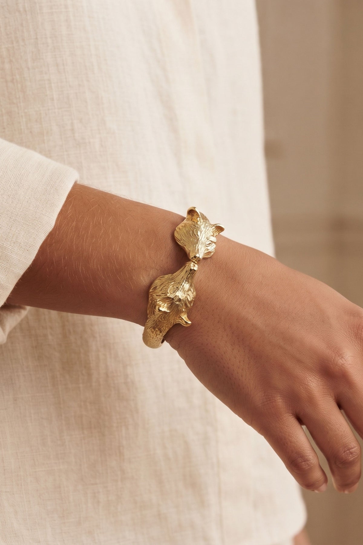 A close-up of a person's wrist wearing a chunky, textured gold cuff bracelet shaped like two overlapping animal heads, likely a fox or rabbit. The person is wearing a light beige linen shirt.
