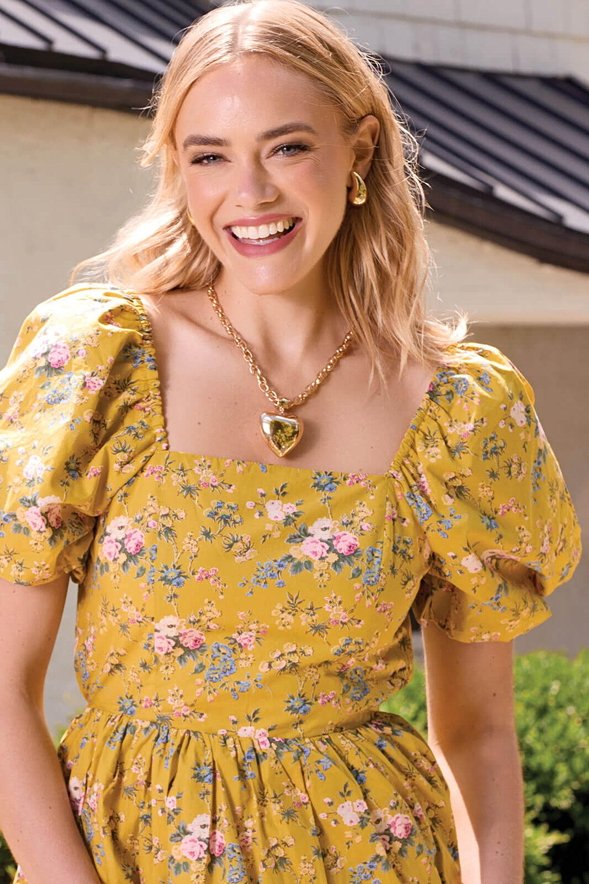 Model in a yellow floral dress wearing the Golden Heart Chain Necklace, the puffed gold heart resting below the neckline.
