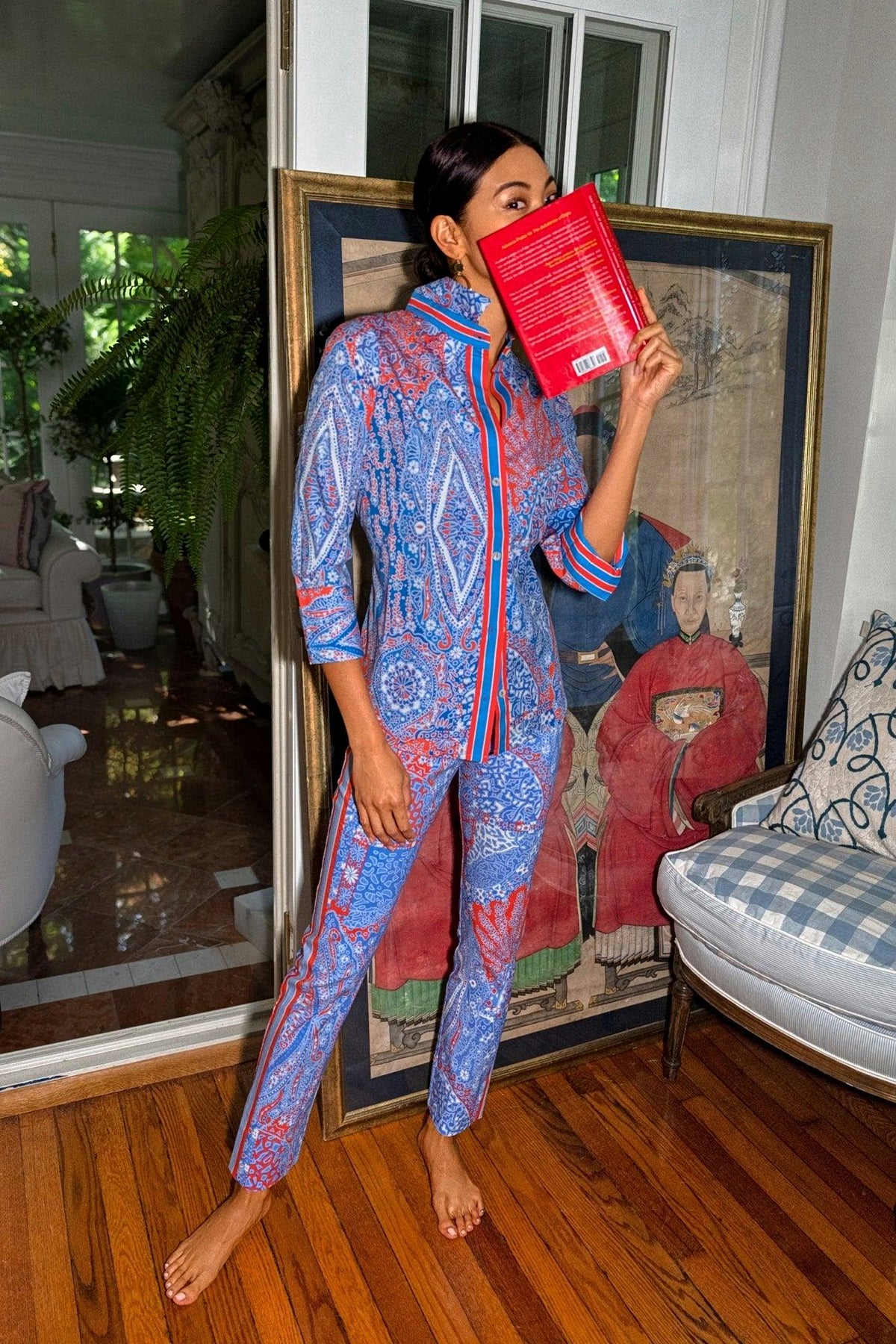 A woman in a matching blue and red paisley print shirt and pull-on pants holds a red book in front of her face.