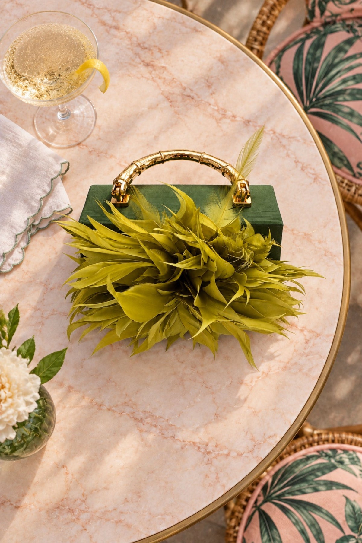 A green rectangular handbag with a gold top handle and a large lime-green feathered or leafy front detail, styled on a round marble-top table with a drink, napkin, and tropical-print cushions nearby.