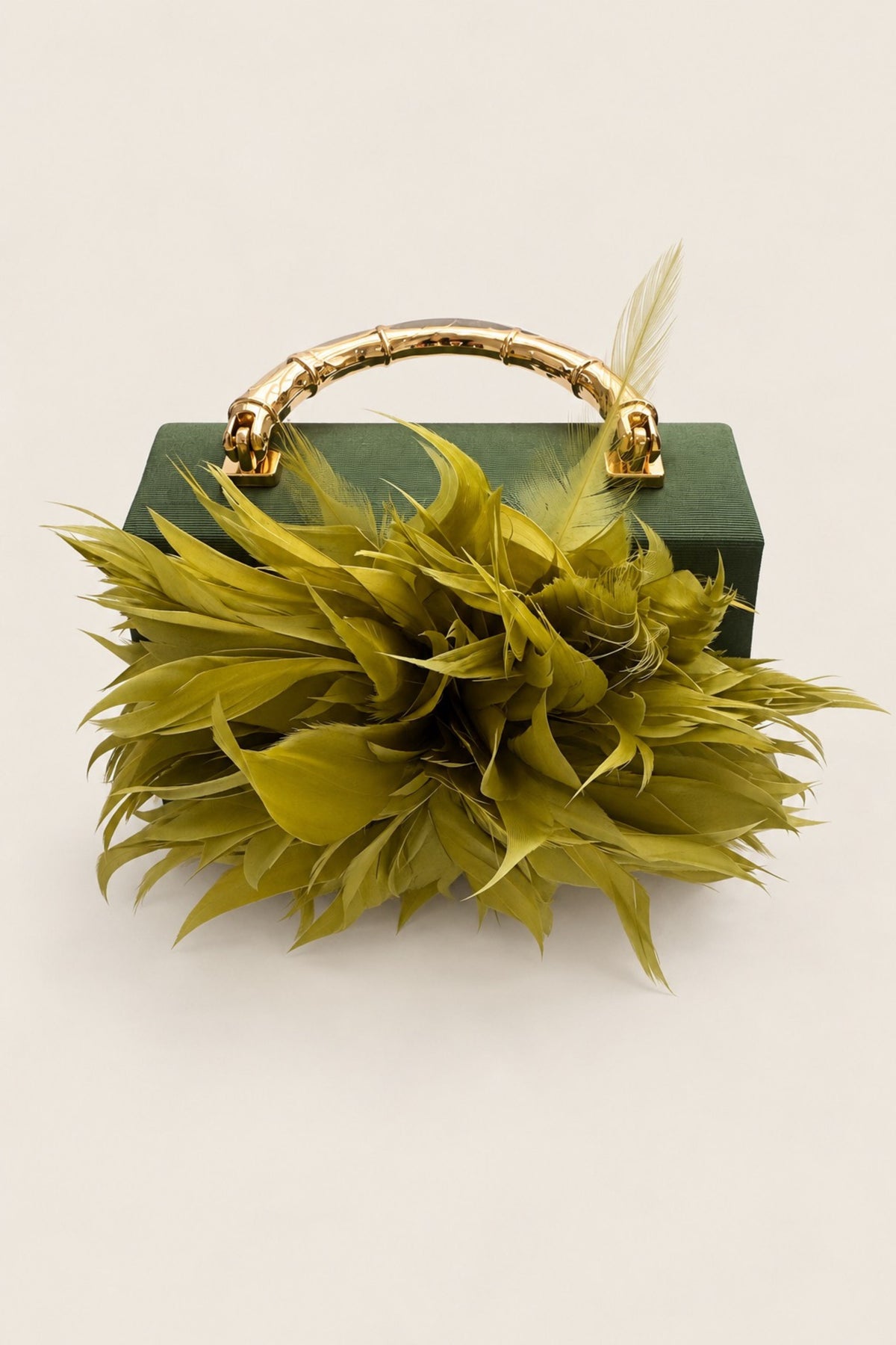 A green rectangular clutch bag with a gold bamboo-style handle, decorated with a large lime-green feathered floral arrangement in front, on a light beige background.