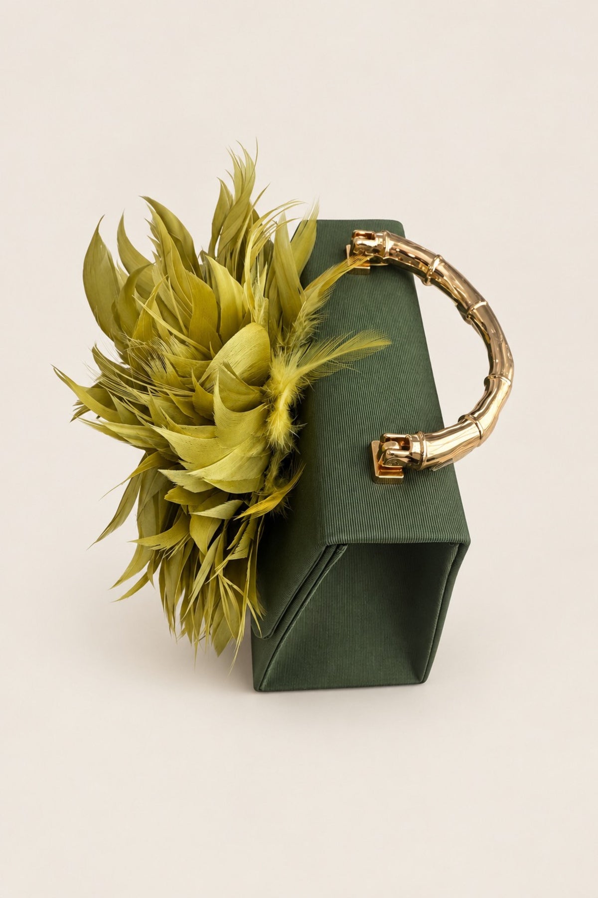 A dark green structured clutch bag with a curved gold handle and a large lime-green feathered floral embellishment on the front, set against a plain light beige background.