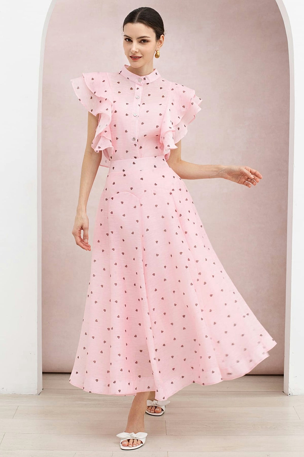A woman in the blush pink heart print organza set: a sleeveless buttoned top with ruffled shoulders and a full maxi skirt.