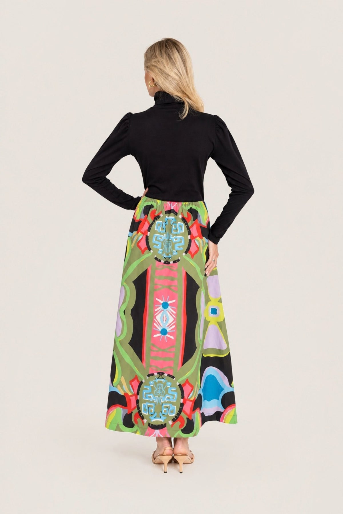 A woman shown from the back wearing a black long-sleeve mock-neck top and a high-waisted, full-length A-line skirt with a vibrant, multicolored geometric and tribal-inspired print. The skirt features shades of green, pink, red, blue, yellow, and black in a bold pattern.