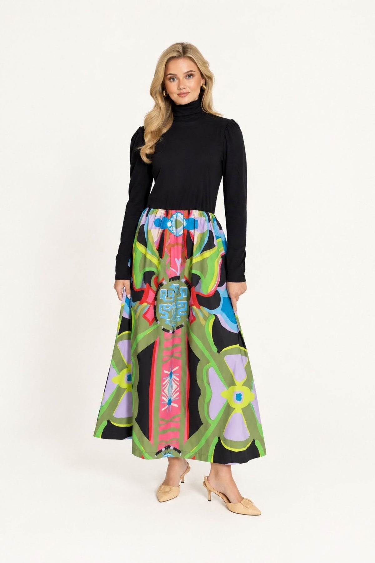 A woman modeling a black long-sleeve mock-neck top paired with a high-waisted, flared midi skirt featuring a vibrant, multicolored geometric and tribal-inspired print on a black base, set against a plain white studio background.