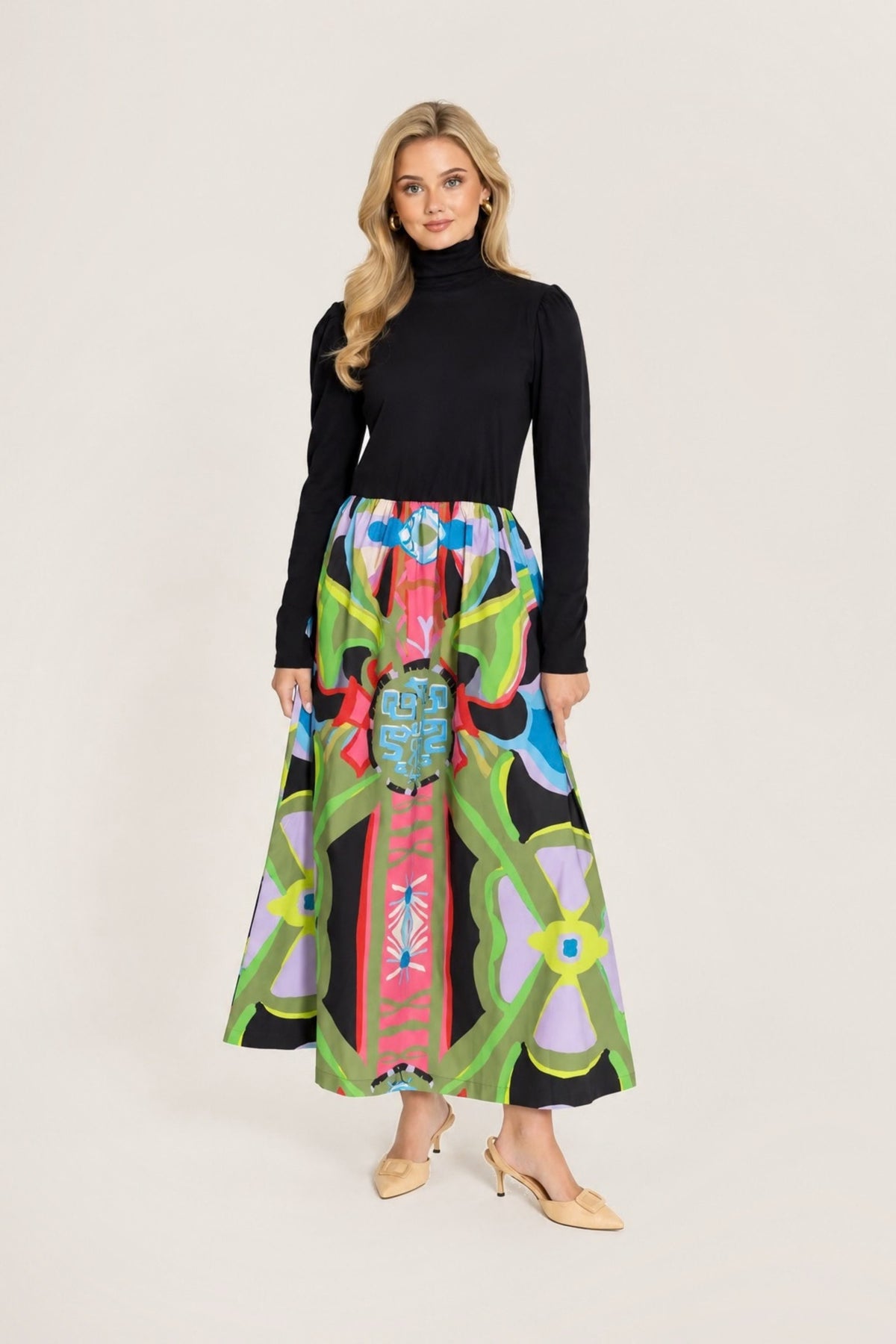 A woman modeling a black long-sleeve mock-neck top paired with a high-waisted, flared midi skirt featuring a vibrant, multicolored geometric and tribal-inspired print on a black base, set against a plain white studio background.