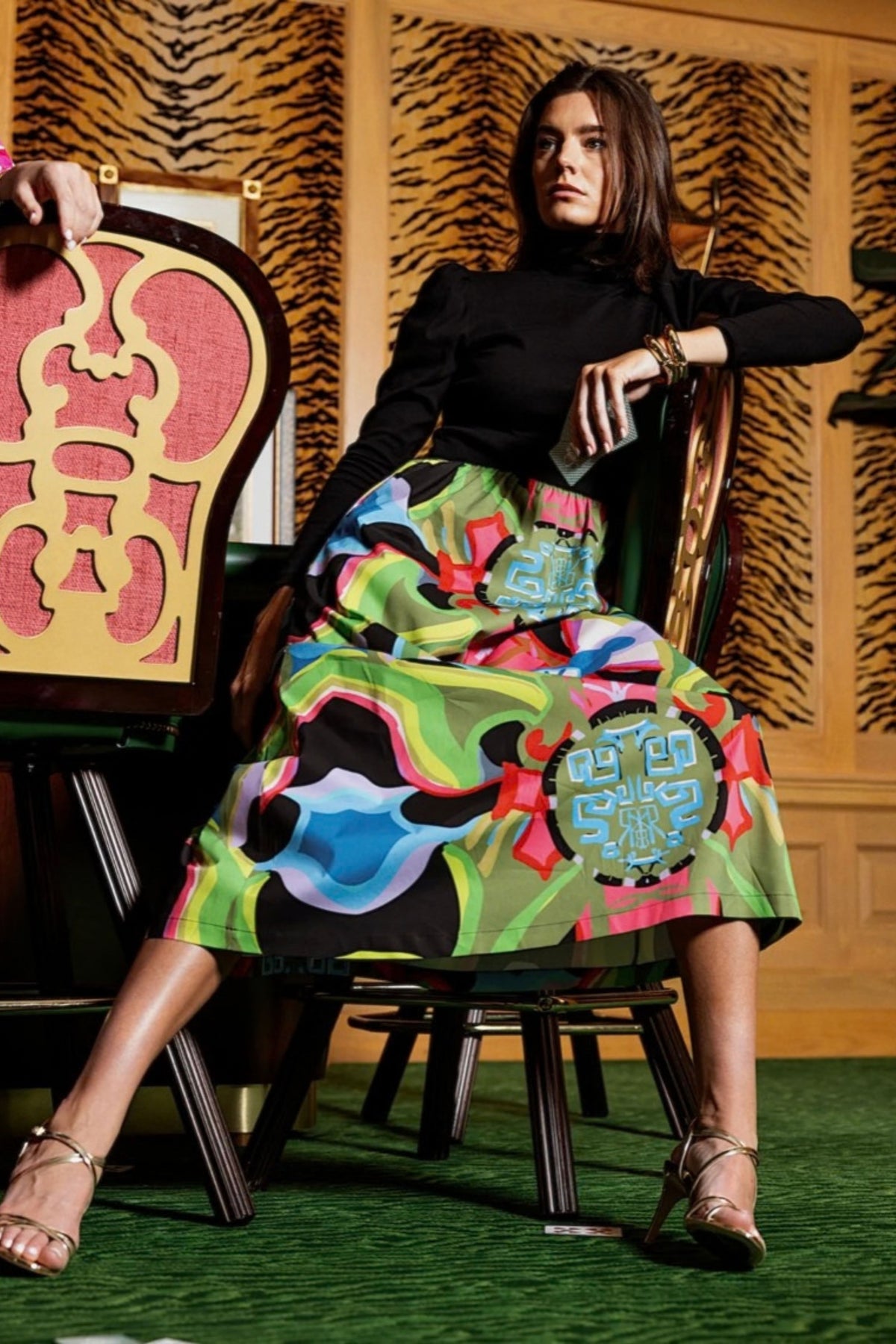 A model wearing a black long-sleeve top and a vibrant, multicolored abstract-patterned midi skirt sits on a wooden chair in a room with tiger-print wallpaper and green carpet.