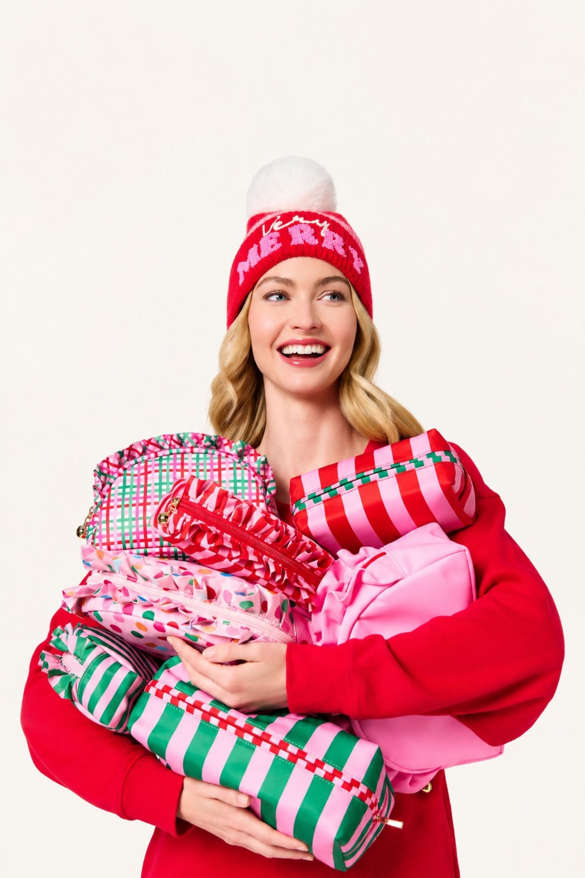 A smiling woman in a red sweater and red knit beanie holds a large pile of Christmas in bright holiday colors against a plain light background.