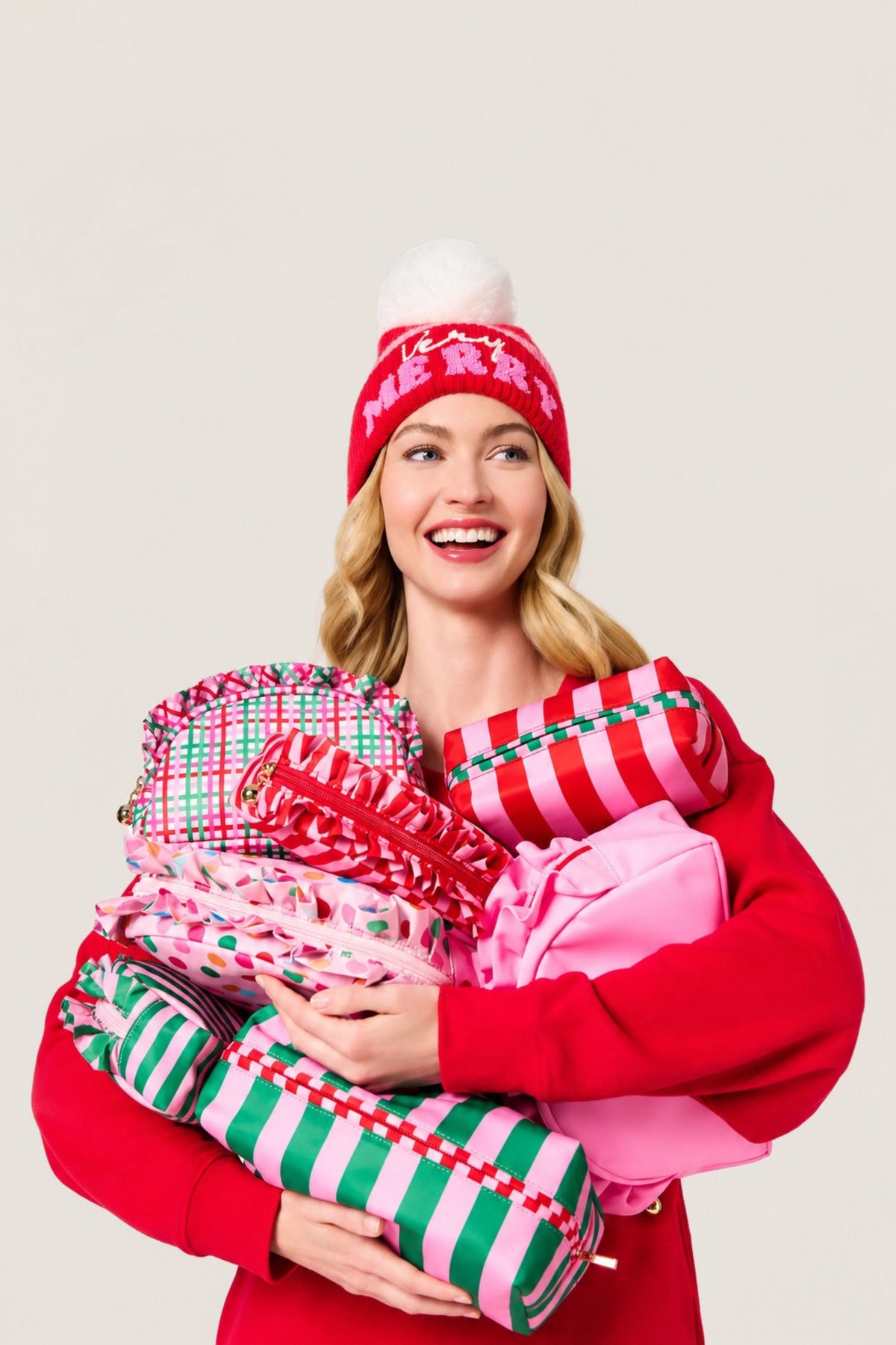 A smiling woman in a red sweater and red knit beanie holds a large pile of Christmas in bright holiday colors against a plain light background.