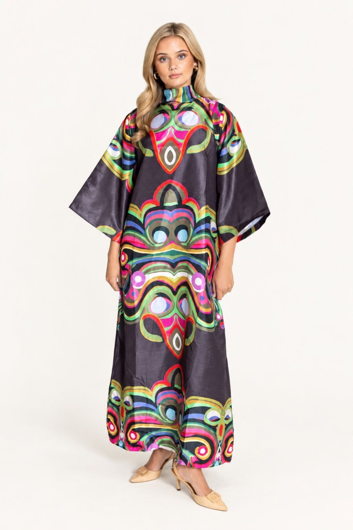 A woman modeling a long, wide-sleeved poncho-style dress with a bold, colorful psychedelic pattern on a dark charcoal background, set against a plain white studio backdrop.