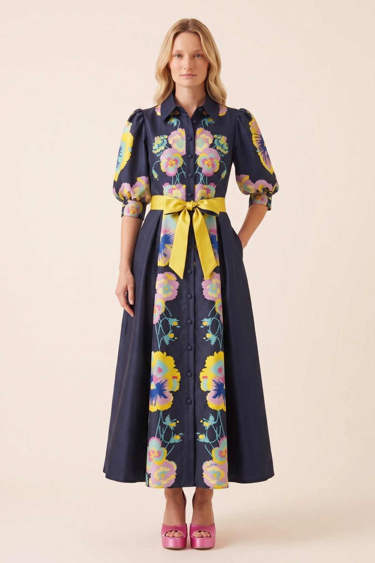 A woman modeling a midnight blue midi dress with a vibrant multicolor floral print, featuring puff sleeves, a button-down front, and a contrasting yellow tie belt at the waist.